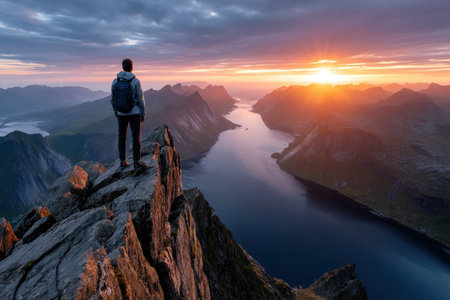 A hiker stands on a mountain peak overlooking a fjord at sunsetの素材