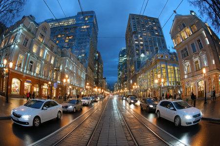 Cars drive down a city street at twilight, past illuminated shops and buildingsの写真素材