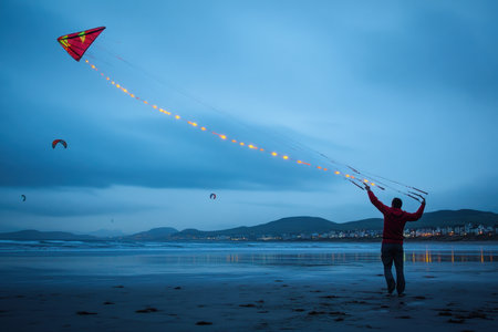 A person stands on the beach flying a brightly lit kite as twilight settles, surrounded by colorful kites.の素材