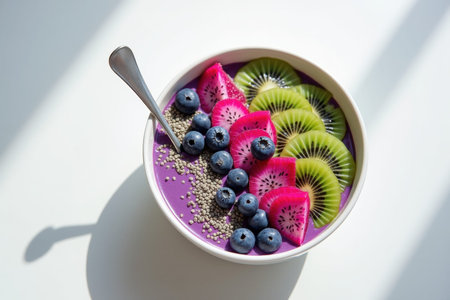 A vibrant purple smoothie bowl is elegantly arranged with fresh kiwi slices, bright pink dragon fruit, ripe blueberries, and small seeds, illuminated by natural light