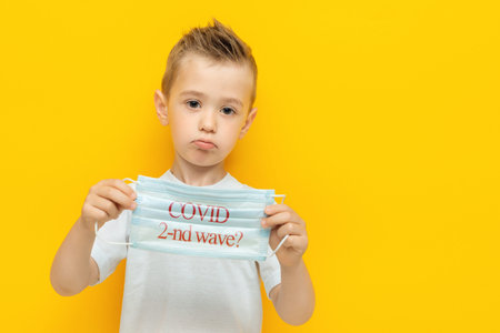 Sad little boy holding a medical mask with the inscription with a question Covid second wave