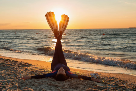 Young woman in a wetsuit lying on the beach stretching up slender legs wearing flippers.