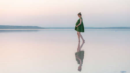 Pregnant girl posing at sunset in the water with a reflection. Amazing sunset over a shallow lake.の写真素材