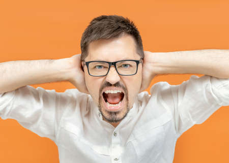 Portrait of emotional nervous bearded man in classic white shirt against an orange background isolated covering ears with hands and screaming. Copy space, bannerの写真素材