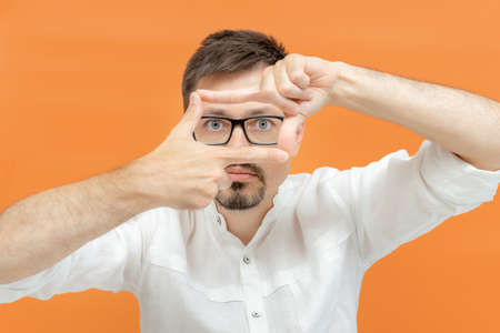 Portrait of attentive curious bearded happy male in white shirt looking at camera, focusing through frame made of fingers. Indoor studio shot isolated on orange background.の写真素材