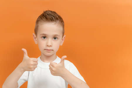 Photo of positive preschool boy show thumb up sign wear with two hands isolated over orange color background. Children studio portrait.の写真素材