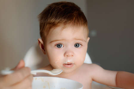 Caucasian baby boy with messy face eating porridge on a high chair. Adorable baby eating food. His mother feeds him with a spoonの写真素材