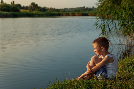 A little happy boy wearing swimming shorts sitting on the shore of a pond at sunset. Concept of freedom and childhood. Child, summer and water. Children's fun in the water. Holidays time.の写真素材
