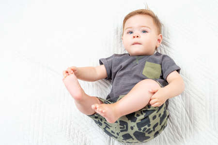 Baby boy in a khaki suit lying on white bed and holding legs. Portrait of adorable toddler on white background, copy space.の写真素材