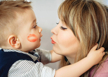 Funny family photo. The one-year-old son hugs his mother tightly and looks at each other. Close-up portrait. Cute happy baby with lipstick mark in his cheek. Family concept.の写真素材