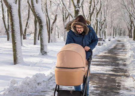 A bearded dad walking with baby carriage in winter park. A man with a baby stroller walks in a winter snow-covered city park in the cold. Caring father admires baby sleeping in strollerの写真素材