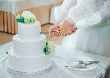 Newlyweds cut the wedding cake. A bride and a groom hands are holding a knife, close up. Beautiful white three-tiered cake decorated with flowersの写真素材