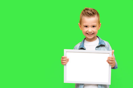 Five year-old boy smiles and holds blank white frame. Happy preschooler child isolated on green...