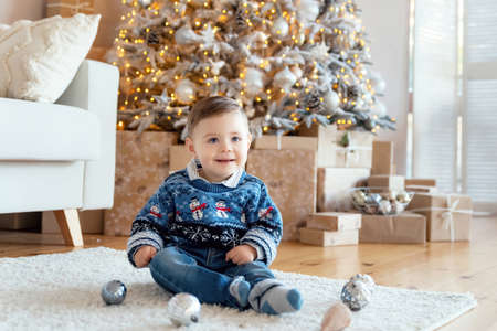 Happy toddler boy wearing knitted Christmas sweater sitting on the floor in Christmas decorated room. Christmas decorations are scattered around the child. New year's eve conceptの写真素材