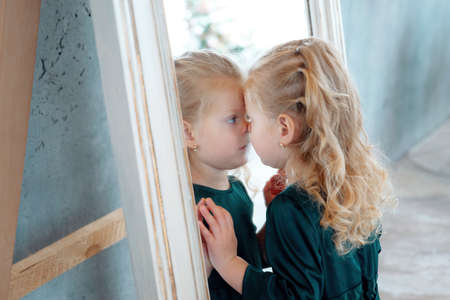 A little cute blonde girl wriggles in the mirror. Funny Caucasian child wearing green dress looks at himself in the mirror.の写真素材