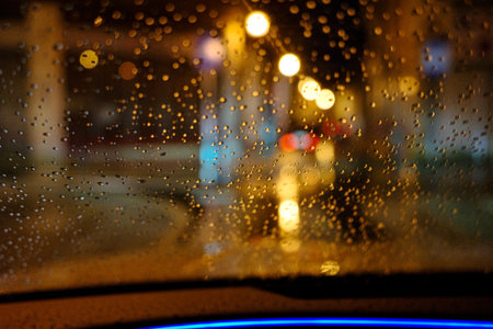 Rain drops on car windshield at night with city lights bokeh, view from inside vehicle during rainy...