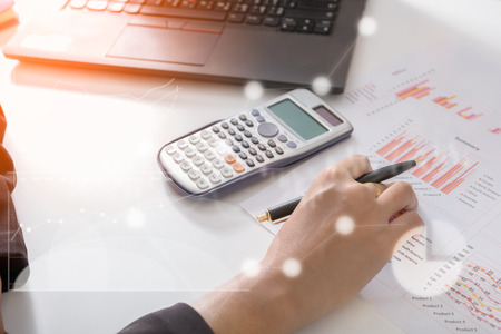 Young finance market analyst working at sunny office on laptop while sitting at white table.Businessman analyze document and the calculator in hands.Graphs and diagram on notebook screenの写真素材