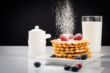 Belgian waffles with raspberries and sieving sugar powder and honey served with jug of milk on a white table.の写真素材