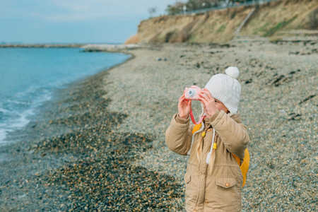a little girl squinting photographs the autumn seascape on a childrens pink camera. the child is...
