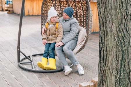grandmother and granddaughter sat down to rest after a walk in a hanging rattan chair outdoors. an adult woman and a child are resting after a walk in autumn, spring on the street in outerwearの写真素材