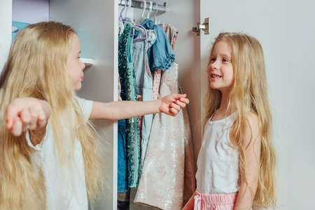 Two little beautiful girls friends sisters play fashion, choose outfits, clothes from a white childrens closet at home indoorsの写真素材