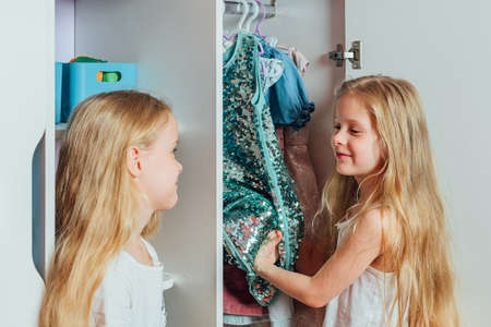 Two little beautiful girls friends sisters play fashion, choose outfits, clothes from a white childrens closet at home indoorsの写真素材
