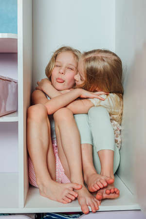 children, sisters, girlfriends, twins play, laugh, have fun, hide at home in the closet with clothesの写真素材