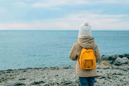 little girl in yellow rubber boots goes to the sea along a rocky beach in spring. view from the back child with a yellow backpack on his back walks. high quality photoの写真素材