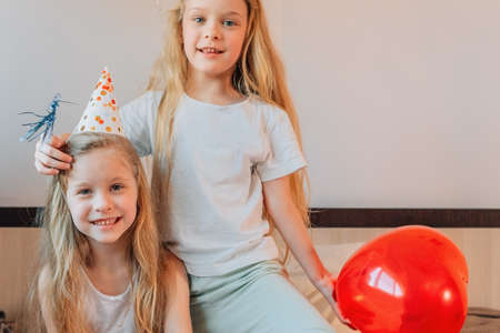 happy children, girls, sisters, twins at home on the bed in the morning on their birthday in festive caps with a red balloon in the form of a heartの写真素材