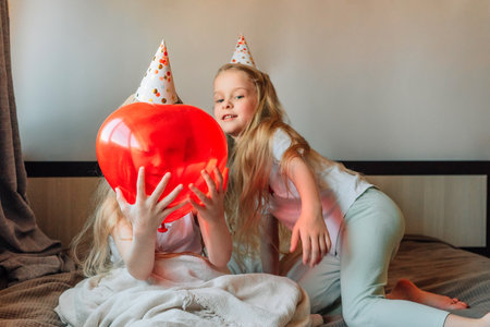happy children, girls, sisters, twins at home on the bed in the morning on their birthday in festive caps with a red balloon in the form of a heartの写真素材