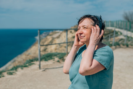 portrait adult woman listens to music in headphones in summer, spring against the backdrop of mountains, enjoy the sea..the pensioner went for a walk in the mountains for health and pleasure, likes to listen to music, books, podcasts in black overhead headphonesの写真素材