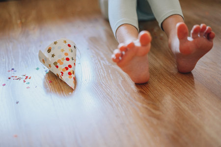 a little girl on her birthday sits on the floor next to scattered confetti from a festive hat. child celebrates his birthday at home during illness, quarantine, isolation. holiday without guestsの写真素材