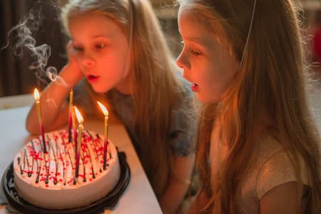 little girls, sisters, twins happily blew out the candles on the birthday cake at home festive cap. child birthday during illness, quarantine, isolationの写真素材