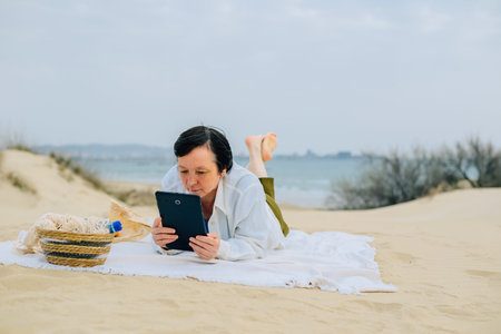 adult woman on the beach by the ocean in spring sits on a picnic, reads a book on a tablet, watches...
