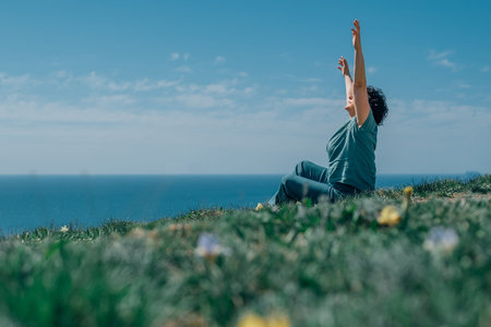 an adult woman sits on a mountain barefoot in a lotus position, raising her hands up to the sky and...