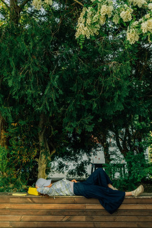 a young woman lies on a wooden bench in summer, covering her face with a baseball capの写真素材