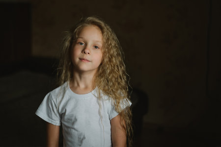 close-up portrait of a little blonde girl with long curly hair with a calm emotion on a dark background, looking at the cameraの写真素材