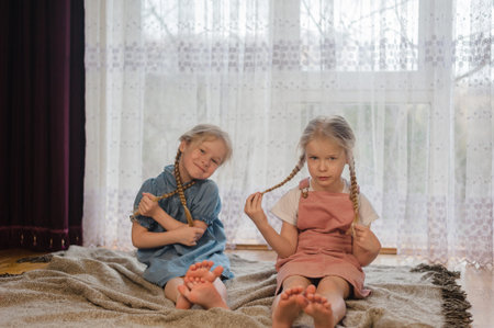 two little sister girls sit at home on the floor and play with their pigtails