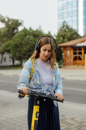portrait of a young woman with an electric scooter in headphones and with a yellow backpack in the summer on a city streetの写真素材