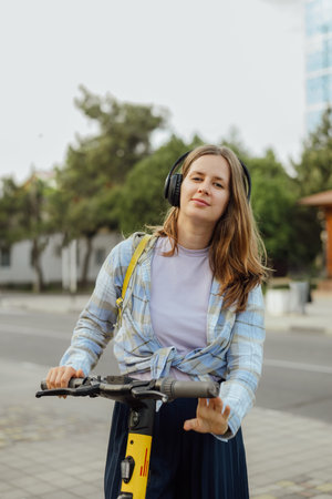 portrait of a young woman with an electric scooter in headphones and with a yellow backpack in the summer on a city streetの写真素材