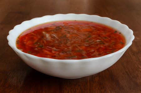 The image shows a white ceramic bowl of tomato soup placed on a wooden surface. The soup is red and appears chunky, with visible bits of vegetables and herbs.の写真素材