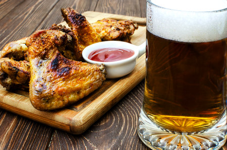 Close-up of a meal consisting of grilled chicken wings, a plate of ketchup and a glass of foamy beer. Chicken wings are golden brown in color and lie on a wooden cutting board.の写真素材