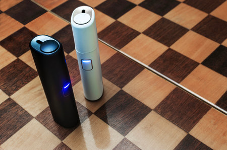 Electronic cigarette technology. Top view of two electric hybrid cigarettes tobacco heating system in white and blue housing on a chessboardの写真素材
