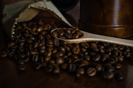Freshly roasted coffee beans spilling from a partially opened brown paper bag onto a wooden surface. A wooden spoon lies among the grainsの写真素材