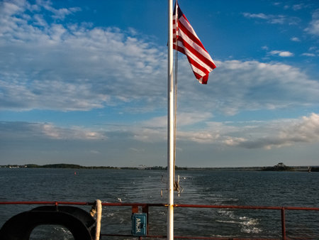 A flag flutters in the wind from a white flagpole on the stern of a tanker. The vessel moves, leaving a foamy trail behind it. The sky is a mixture of blue and white with scattered clouds stretching across the horizon.の写真素材