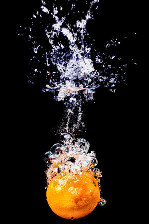 The moment an orange is dipped into water, creating a stunning splash effect. The bright orange fruit contrasts sharply with the dark background, highlighting the dynamic movement of water droplets and air bubbles.の写真素材