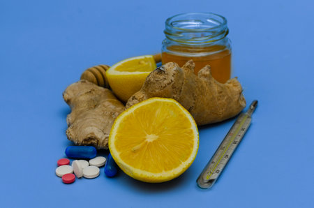 A composition of natural remedies and pharmaceutical medications for treating cold and flu symptoms. The image features fresh lemon, ginger root, honey in a glass jar, a thermometer, and various pills on a blue background, symbolizing the contrast between traditional and modern medicine.の写真素材