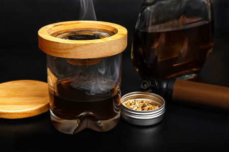 A stylish glass of whiskey infused with a wooden smoker, releasing delicate smoke, accompanied by a bottle, wood chips, and a modern smoking tool on a dark background.の写真素材