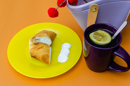 A vibrant still life featuring a pancake filled with cream on a yellow plate, paired with a purple mug of tea with a lemon slice, set against an orange background with decorative pom-poms.の写真素材