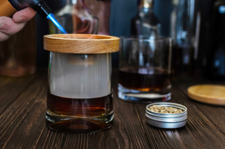 A close-up shot of a cocktail being smoked using a handheld torch and wood chips. The smoke is captured inside a glass with a wooden topper, showing the process of adding a smoky flavor to a drink.の写真素材